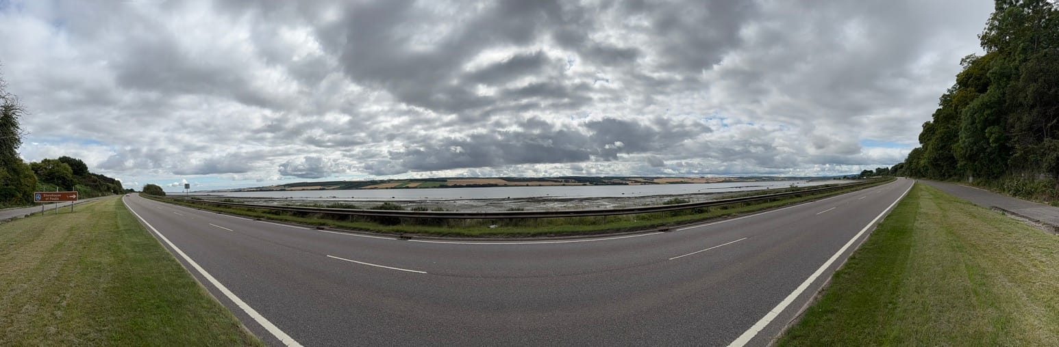 Panoramic view of the Beauly Firth beside a single carriageway under dramatic clouds.
