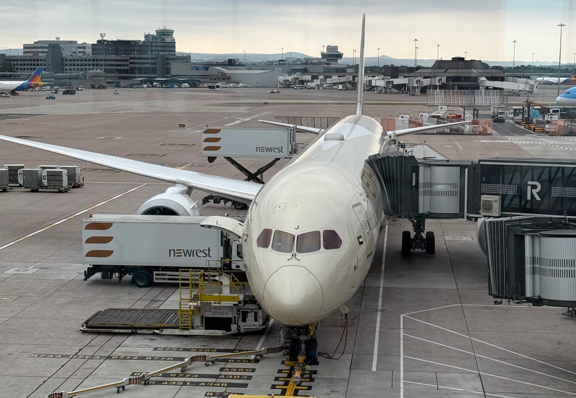 Etihad Airways Boeing 787 Dreamliner parked at a gate at Manchester Airport, with catering vehicles and an air bridge attached before an international departure.