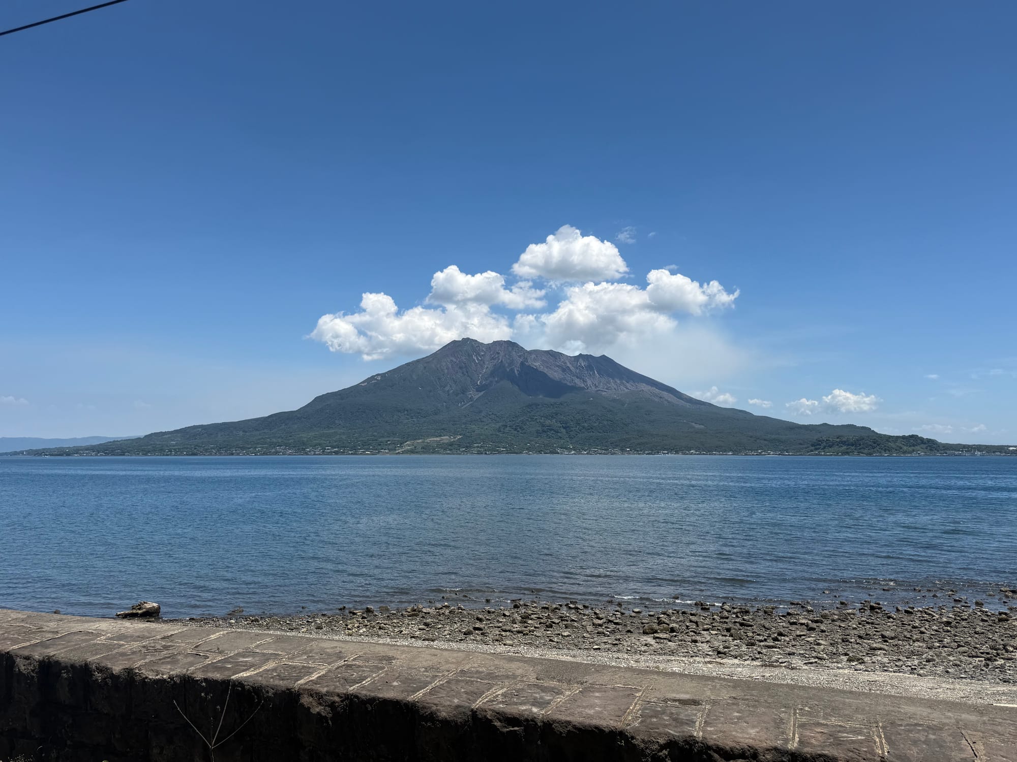 Sakurajima volcano rises across Kagoshima Bay beneath a bright blue sky, with calm water, a rocky shoreline and a cluster of white clouds above the summit.