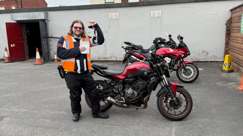 “Person in motorcycle gear standing beside a red motorcycle, holding up a certificate in a parking area.”