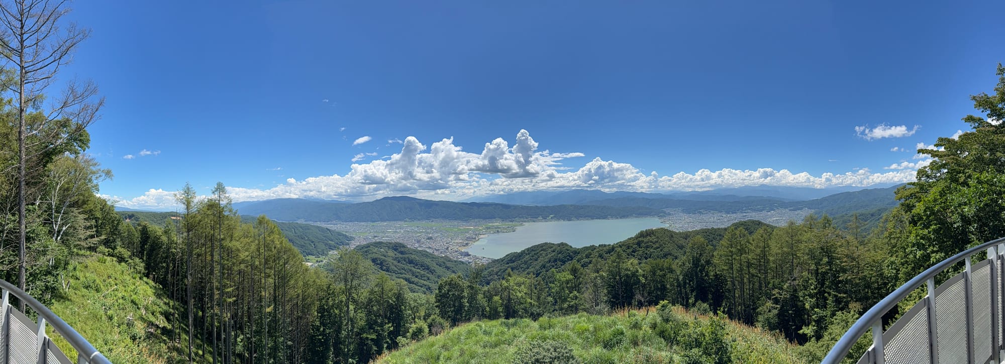 Panoramic view of Lake Suwa from a mountain lookout, showing the lake, surrounding towns and forested hills beneath a clear blue sky with towering summer clouds.