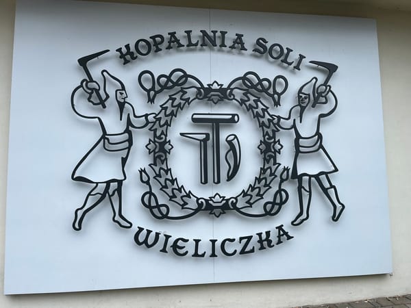 Sign for the Wieliczka Salt Mine featuring two miners holding tools on either side of the mine's emblem, mounted on a white wall.