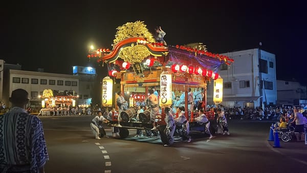 Illuminated festival float decorated with lanterns and carvings, paraded through the streets of Kazuno at night, accompanied by musicians and participants in traditional dress.