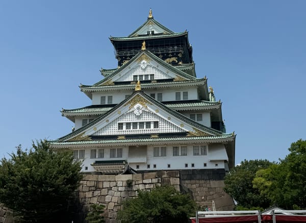 Osaka Castle in central Osaka, featuring its distinctive white walls, green roofs and gold ornamentation rising above stone ramparts beneath a clear blue sky.