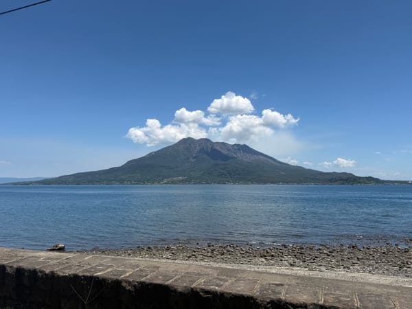 Sakurajima volcano rises across Kagoshima Bay beneath a bright blue sky, with calm water, a rocky shoreline and a cluster of white clouds above the summit.