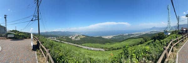 Panoramic view from a hillside viewpoint above Beppu, overlooking green mountains, the city, Beppu Bay and the distant coastline beneath a clear summer sky.