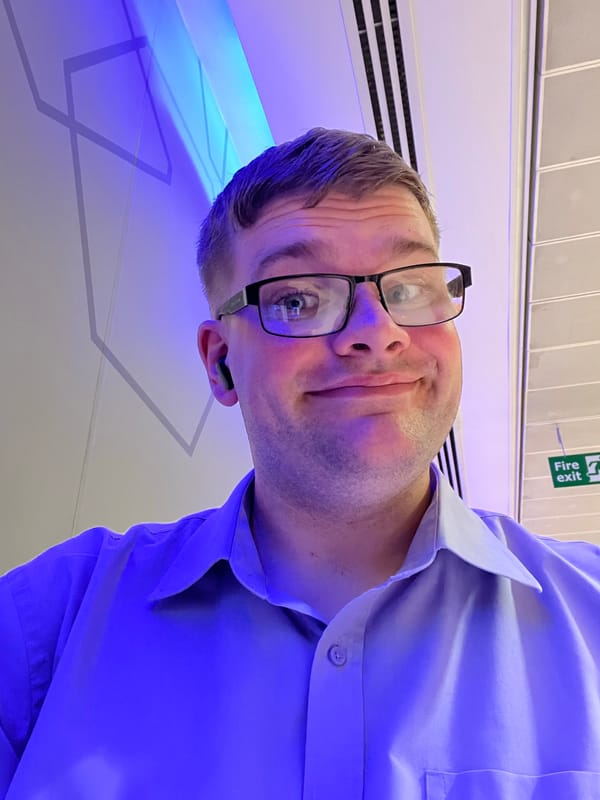 Person wearing glasses, a light blue shirt and wireless earbuds takes a selfie indoors under blue lighting, with geometric wall designs and a fire exit sign visible behind.