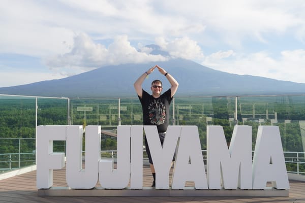 Visitor standing behind large FUJIYAMA letters at Fujiyama Tower, with Mount Fuji rising in the background above forests and scattered clouds on a clear day.